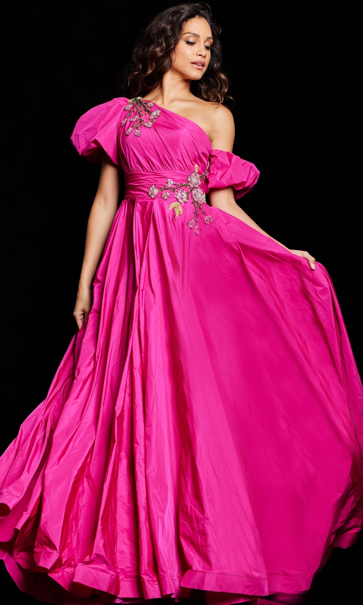 A person stands against a black background wearing the Long Prom Dress 36872 by Jovani—a bright pink, one-shoulder taffeta gown featuring floral embroidery and puffed sleeves.